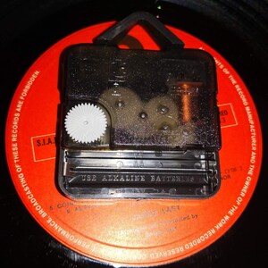May include: A black clock mechanism with a white gear is mounted on a red vinyl record. The mechanism has a battery compartment with the text "USE ALKALINE BATTERIES". The record has printed text around the edge.