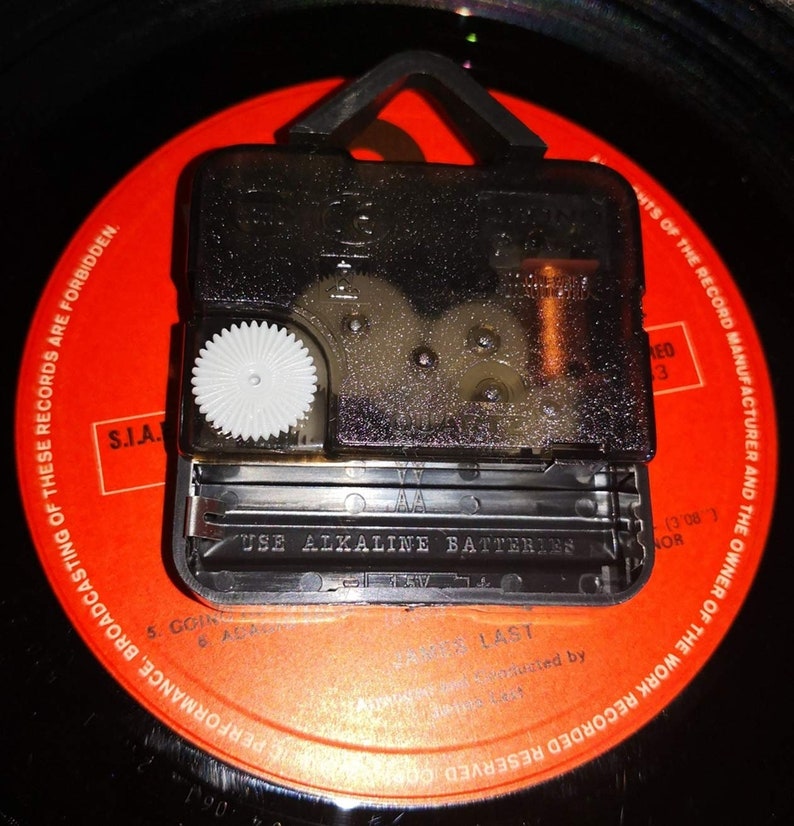 May include: A black clock mechanism with a white gear is mounted on a red vinyl record. The mechanism has a battery compartment with the text "USE ALKALINE BATTERIES". The record has printed text around the edge.