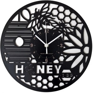 Op de afbeelding: Een zwart-witte decoratieve wandklok met een bijen- en honingraatontwerp. De wijzerplaat is zwart met witte wijzers en het woord "HONEY" wordt weergegeven. De klok is rond en heeft een diameter van ongeveer 30 cm.