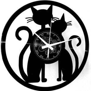 May include: Black cat-themed wall clock. The clock features two cat silhouettes within a circular frame. The clock face has Roman numerals and gear-like details. The hands are white.