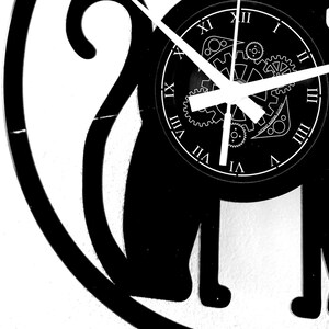 May include: Black and white cat-shaped wall clock. The clock face features Roman numerals and a gear design. The clock hands are white, and the text "INSTANT KARMA CLOCKS" is visible on the clock face. The clock is mounted on a white wall.