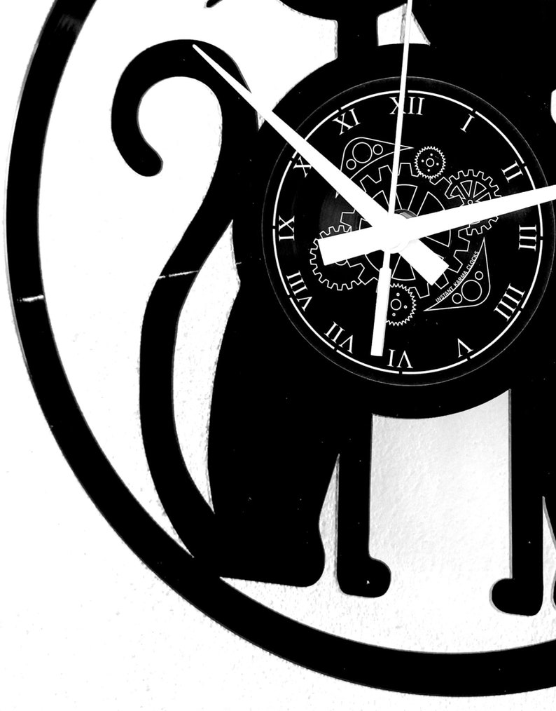May include: Black and white cat-shaped wall clock. The clock face features Roman numerals and a gear design. The clock hands are white, and the text "INSTANT KARMA CLOCKS" is visible on the clock face. The clock is mounted on a white wall.