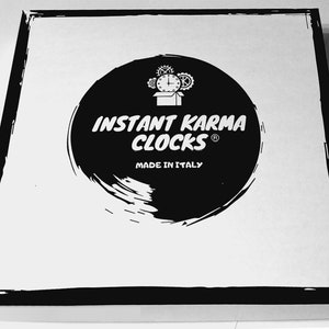 May include: A square, white box with a black border. The box features a large, black circular design with the words "INSTANT KARMA CLOCKS" in white, along with a clock graphic and "MADE IN ITALY".