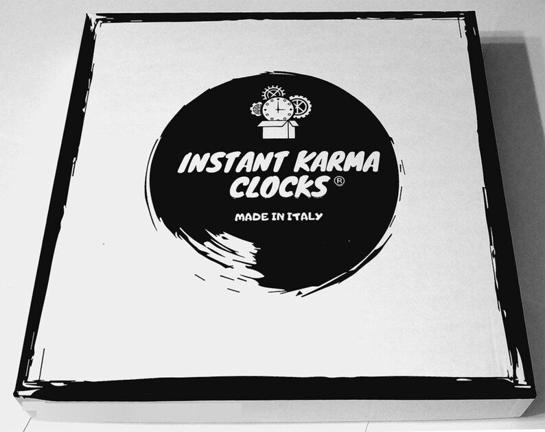 May include: A square, white box with a black border. The box features a large, black circular design with the words "INSTANT KARMA CLOCKS" in white, along with a clock graphic and "MADE IN ITALY".
