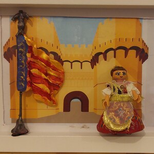 May include: A framed miniature scene featuring a woman in a red and gold dress standing in front of a yellow castle with a red and yellow flag.