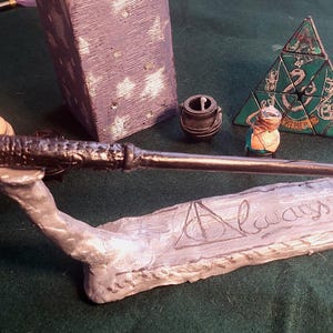 Puede incluir: Una varita oscura descansa sobre una base pintada de plata con la palabra "Always" y un símbolo triangular. Otros elementos incluyen un pequeño caldero, un rompecabezas triangular verde y blanco y una pequeña figura. Los elementos están sobre una superficie verde.