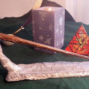 Peut inclure: Une baguette marron repose sur un support argenté avec l'inscription "Always". Une bougie violette et un puzzle pyramidal rouge et jaune sont également présents. Les objets sont disposés sur une surface verte.