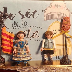 May include: A framed diorama with two Playmobil figures in traditional dress, a flag, and text. The figures are a woman in a blue and gold dress and a boy in a vest and shorts. The flag is orange and yellow. The text reads "Jo sóc de la festa."