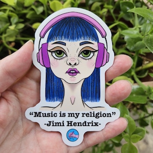 Op de afbeelding: Een sticker met een gestileerde illustratie van een persoon met blauw haar en groene ogen die een roze koptelefoon draagt. De sticker bevat de quote "Music is my religion" van Jimi Hendrix.