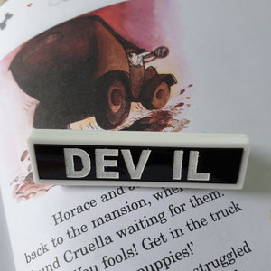 May include: A rectangular badge with the word "DEVIL" in silver lettering on a black base, edged with white. The badge is placed on a book page with an illustration of a vintage car.