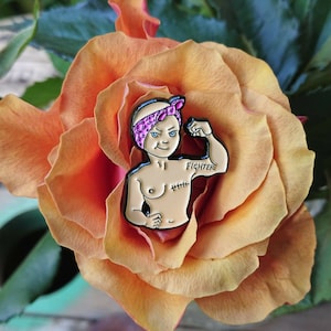 May include: A colourful enamel pin depicting a strong woman with a pink headscarf and the word "Fighter" tattooed on her arm. The pin has a black outline, set against an orange rose.