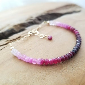 May include: A delicate gold chain bracelet with a gradient of pink and red gemstones. The bracelet features a small heart-shaped charm.