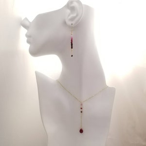 May include: A gold necklace and earrings set with a teardrop-shaped red gemstone and smaller pink and red gemstones. The necklace and earrings are on a white mannequin.