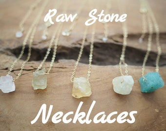 Puede incluir: Cinco collares de oro con colgantes de piedra en bruto. Las piedras son de varios tonos de verde, blanco y amarillo. Los collares se muestran sobre una superficie de madera. Collares de piedra en bruto.