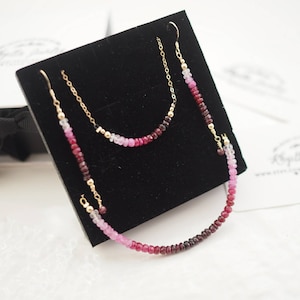 May include: A set of three gold necklaces with pink, red, and purple gemstone beads. The necklaces are displayed on a black velvet background.
