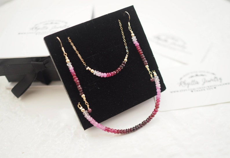 May include: A set of three gold necklaces with pink, red, and purple gemstone beads. The necklaces are displayed on a black velvet background.