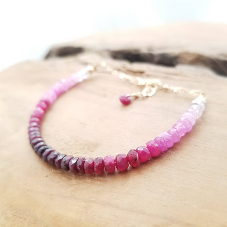 May include: A delicate bracelet made with a gradient of pink and red gemstones. The bracelet is made with small, faceted beads that are strung on a gold chain.