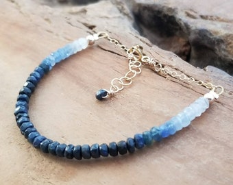 Puede incluir: Un brazalete de piedras preciosas de zafiro azul con un degradado de cuentas azules, que van del azul oscuro al azul claro. El brazalete tiene un cierre dorado y está hecho con cuentas pequeñas y facetadas.