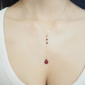 May include: A gold chain necklace with a teardrop-shaped red gemstone pendant and small red and pink gemstones along the chain.