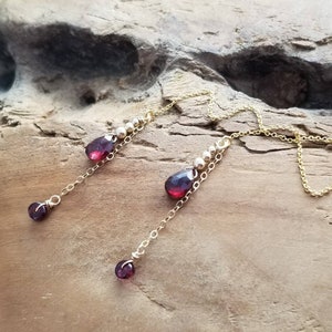 Puede incluir: Un par de aretes largos y delicados con una cadena dorada y gotas de piedras preciosas rojas. Los aretes presentan pequeñas cuentas doradas a lo largo de la cadena.