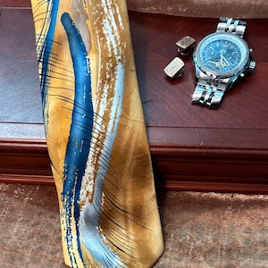 Pode incluir: Uma gravata com fundo dourado, uma pincelada azul proeminente, destaques brancos e linhas pretas finas. Ao lado da gravata, encontram-se um relógio de pulso analógico prateado com mostrador azul-escuro e dois pequenos botões de punho retangulares.