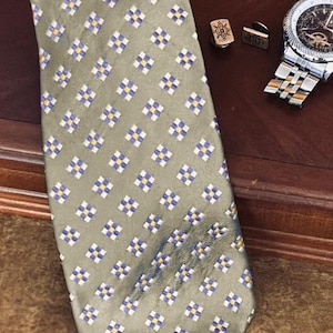 May include: An olive green necktie featuring a repeating pattern of small white, blue, and yellow squares. Beside the tie are two rectangular metal cufflinks with engraved details and a silver-tone analog wristwatch with a metallic link band.