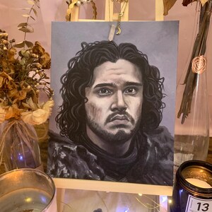 Puede incluir: Un retrato de un hombre con cabello rizado oscuro y barba, sobre un pequeño caballete de madera. La obra presenta una expresión sombría y está realizada en tonos grises y marrones. El fondo es gris apagado.