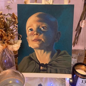 May include: An oil painting of a young child with a bald head and a green hooded cloak. The child is looking upwards with a soft, contemplative expression. The painting is displayed on a small wooden easel.