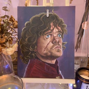 Puede incluir: Una pintura de retrato de un hombre con cabello castaño rizado y ojos azules, sobre un fondo púrpura. El hombre tiene una cicatriz en la cara y viste una prenda roja. La obra de arte se exhibe en un pequeño caballete de madera.