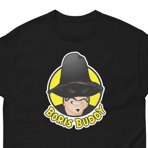 Peut inclure: T-shirt noir avec un graphique circulaire jaune d'un homme portant un chapeau noir et un masque. Le texte "Boris Buddy" est écrit en lettres blanches autour du cercle.