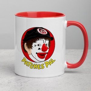 Op de afbeelding: Een witte keramische mok met een rode rand en handvat. De mok heeft een cartoonclown met een rode neus en een rood en wit gezicht. De clown draagt ​​een zwarte hoed en de tekst "Patches Pal" staat in geel onder de clown.