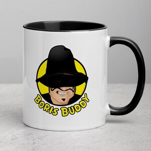 Peut inclure: Une tasse en céramique blanche avec un bord et une poignée noirs. La tasse présente une illustration de dessin animé d'une personne portant un chapeau noir et des lunettes de soleil avec le texte "Boris Buddy" en lettres jaunes.