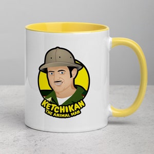 May include: White ceramic mug with a yellow rim and handle. The mug features a cartoon illustration of a man wearing a green shirt and a brown hat with the text "Ketchikan The Animal Man".