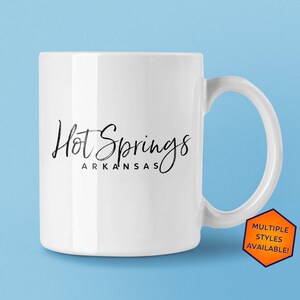 May include: White ceramic mug with a black handwritten design that says "Hot Springs Arkansas".