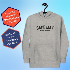 May include: Gray hooded sweatshirt with black text that reads "CAPE MAY NEW JERSEY". The sweatshirt is hanging on a hanger. There are two hexagonal shapes with text in the background. The first hexagon is orange and says "CHOOSE FROM 15 EMBROIDERY COLORS!" The second hexagon is blue and says "CHOOSE FROM 12 HOODIE COLORS!"