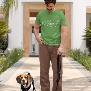 May include: A man wearing a green t-shirt with the text "Wildwood New Jersey" in white lettering walks with a beagle dog on a leash.