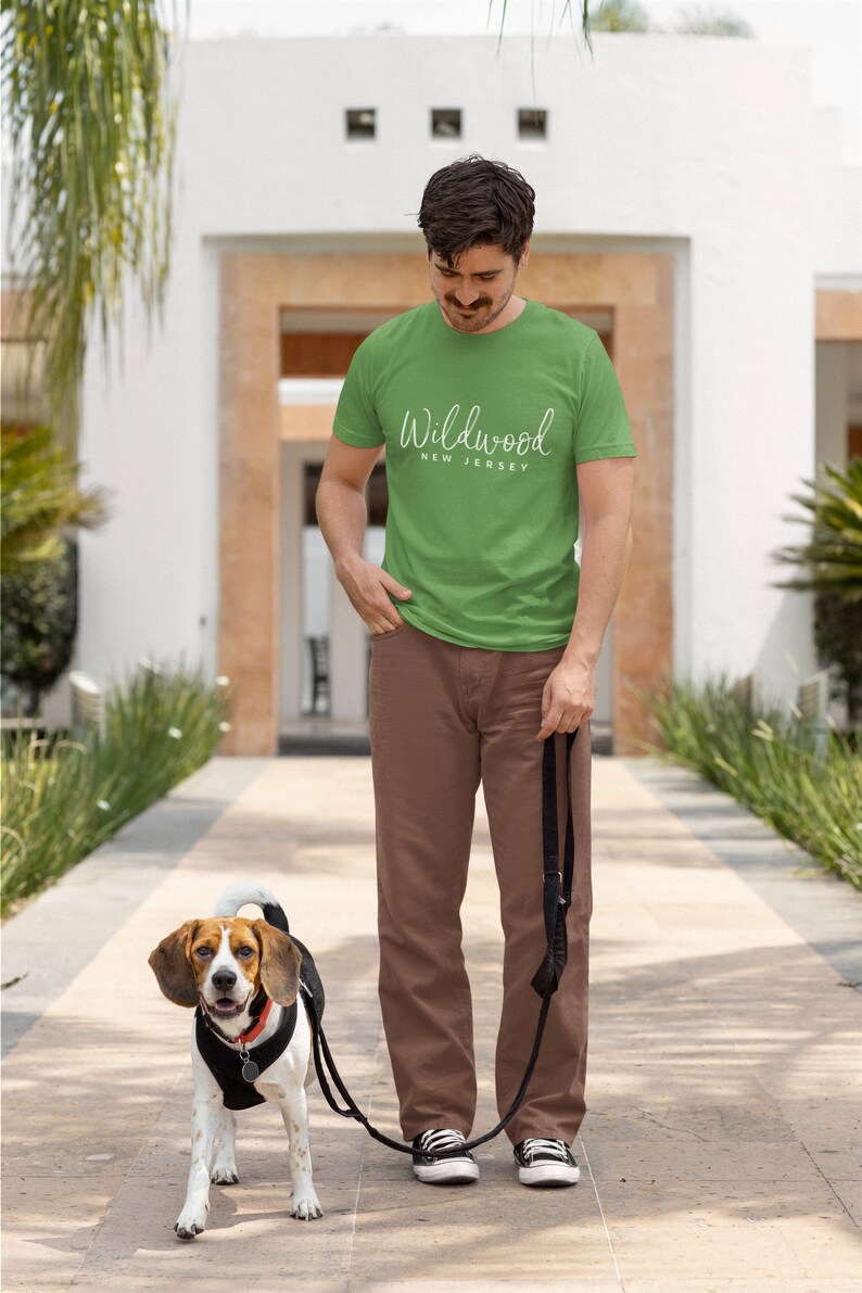 May include: A man wearing a green t-shirt with the text "Wildwood New Jersey" in white lettering walks with a beagle dog on a leash.
