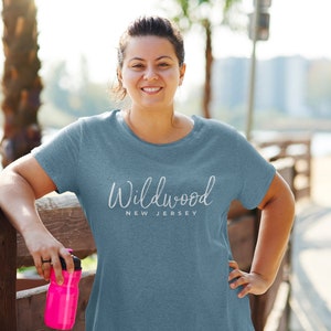 May include: A woman wearing a light blue t-shirt with the text "Wildwood New Jersey" in white. She is standing next to a wooden fence and holding a pink water bottle.