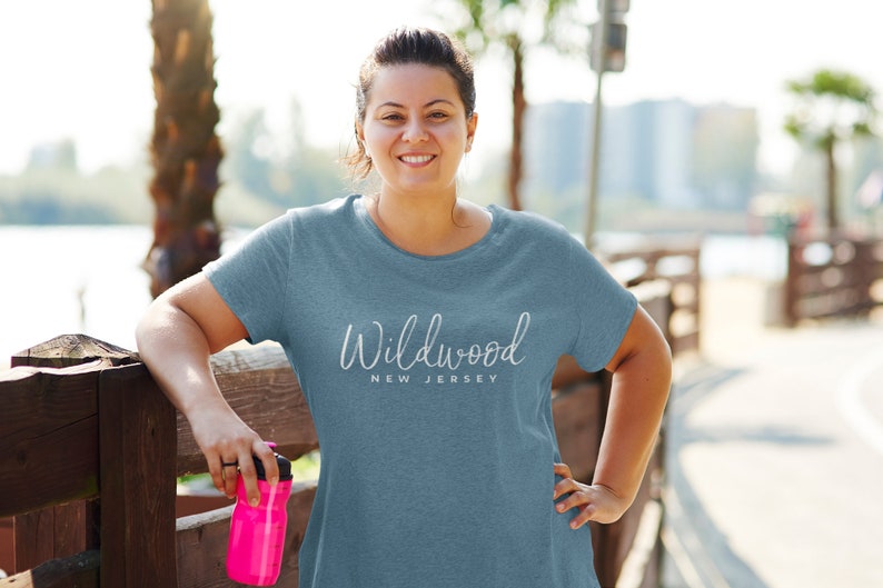 May include: A woman wearing a light blue t-shirt with the text "Wildwood New Jersey" in white. She is standing next to a wooden fence and holding a pink water bottle.