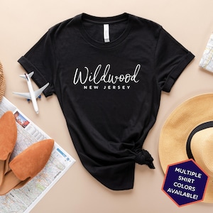 May include: Black t-shirt with white text that reads "Wildwood New Jersey". The shirt is knotted and laid on a beige surface with a straw hat, a pair of brown shoes, and a map.