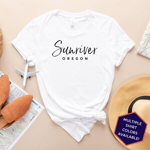 May include: White t-shirt with black text that reads "Sunriver Oregon". The shirt is photographed on a light brown background with a straw hat, a map, and a toy airplane.