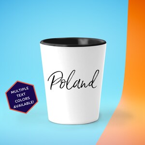 May include: White shot glass with a black rim. The glass has the word "Poland" written in black cursive script.