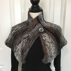 May include: A hand-knitted, multi-colored shawl with gray, brown, and white yarn. The shawl is fastened with two large, round buttons. The garment is displayed on a mannequin, showcasing its textured knit design.