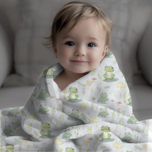 May include: A baby wrapped in a white and green baby blanket with a frog pattern. The blanket has a white background with green frogs, leaves, and flowers.