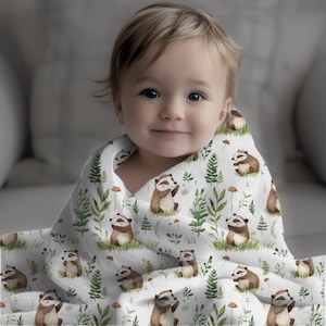 Könnte beinhalten: Eine weiße Babydecke mit einem sich wiederholenden Muster aus braunen und weißen Dachsen, grünen Blättern und kleinen roten Pilzen. Ein Baby ist in die Decke gewickelt und schaut direkt in die Kamera.