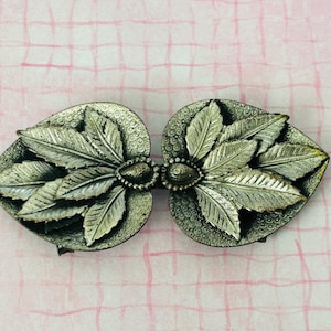 May include: Silver-toned metal clasp with a leaf design. The clasp is composed of two heart-shaped halves, each adorned with detailed leaf patterns and a central oval embellishment. The clasp is set against a pink and white checkered background.