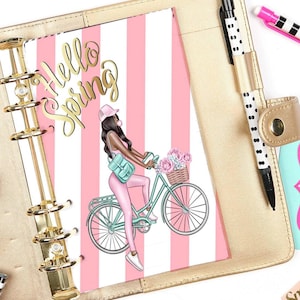 Puede incluir: Una agenda rayada rosa y blanca con un anillo de encuadernación dorado. La agenda tiene una inscripción dorada que dice "Hello Spring" y una ilustración colorida de una mujer montando una bicicleta con una cesta de flores.