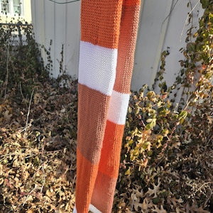 Puede incluir: Una bufanda de punto con un dise&ntilde;o de bloques de color. La bufanda presenta rayas alternas de color naranja, blanco y marr&oacute;n. La bufanda est&aacute; colgada afuera en un alambre. La bufanda est&aacute; hecha de un material suave y c&aacute;lido.