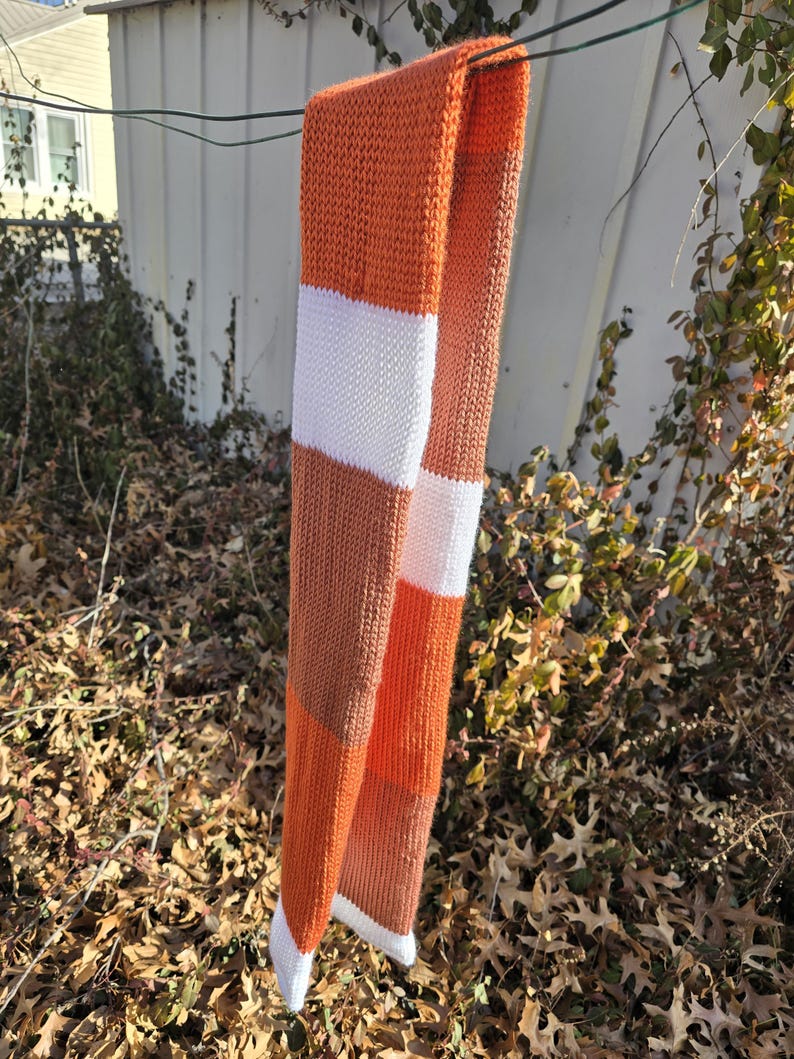 Puede incluir: Una bufanda de punto con un dise&ntilde;o de bloques de color. La bufanda presenta rayas alternas de color naranja, blanco y marr&oacute;n. La bufanda est&aacute; colgada afuera en un alambre. La bufanda est&aacute; hecha de un material suave y c&aacute;lido.
