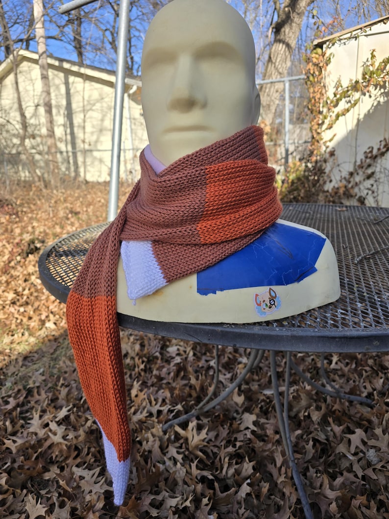 Puede incluir: Una bufanda de punto en tonos marr&oacute;n, naranja y blanco est&aacute; drapeada sobre una cabeza de maniqu&iacute;. La bufanda presenta un dise&ntilde;o de bloques de color con una mezcla de secciones lisas y rayadas. La bufanda se exhibe al aire libre sobre una mesa de metal.